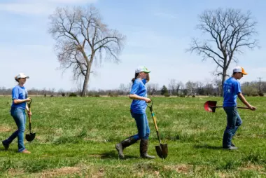 Three people carry shovels across a field to participate in a spring tree planting event in Wardensville, West Virginia.